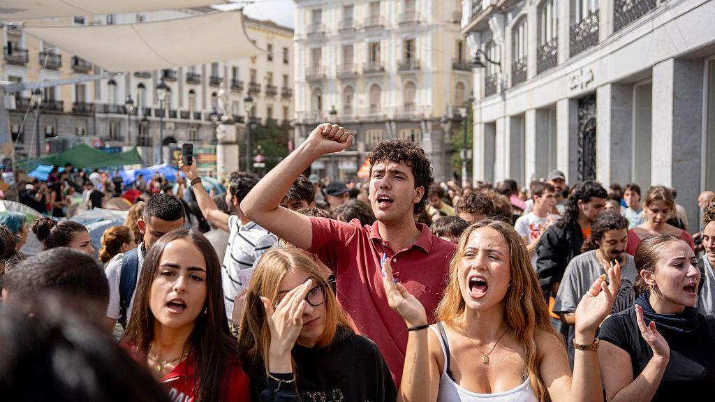 Four women and a man shout and raise their arms while protesting at Puerta del Sol in Madrid. Crowds of other people stand around them.