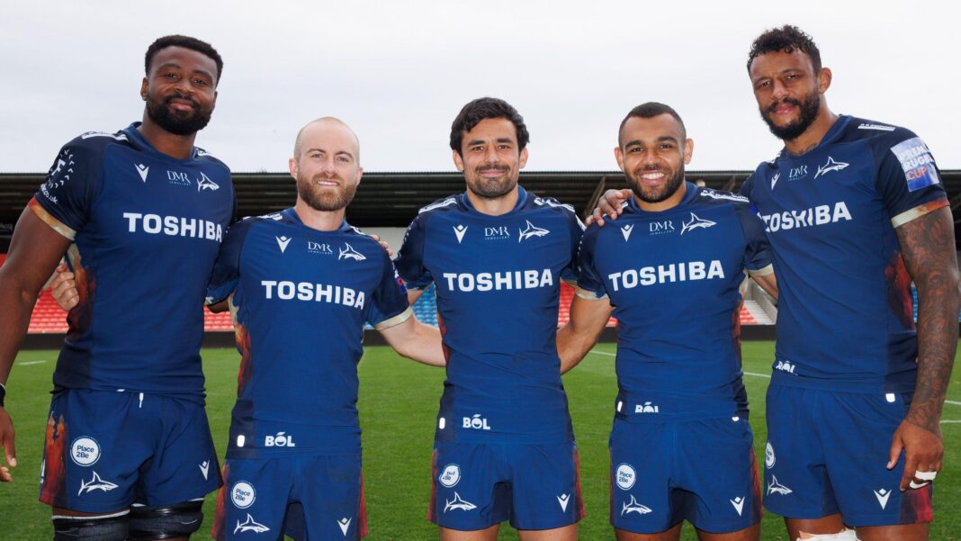 Christ Tshiunza, Xavier Roe, Alex Lozowski, Joe Marchant and Courtney Lawes stand in a line with their arms around each other, looking straight ahead. They are all wearing blue Sale kit having just played a Prem Rugby Cup game