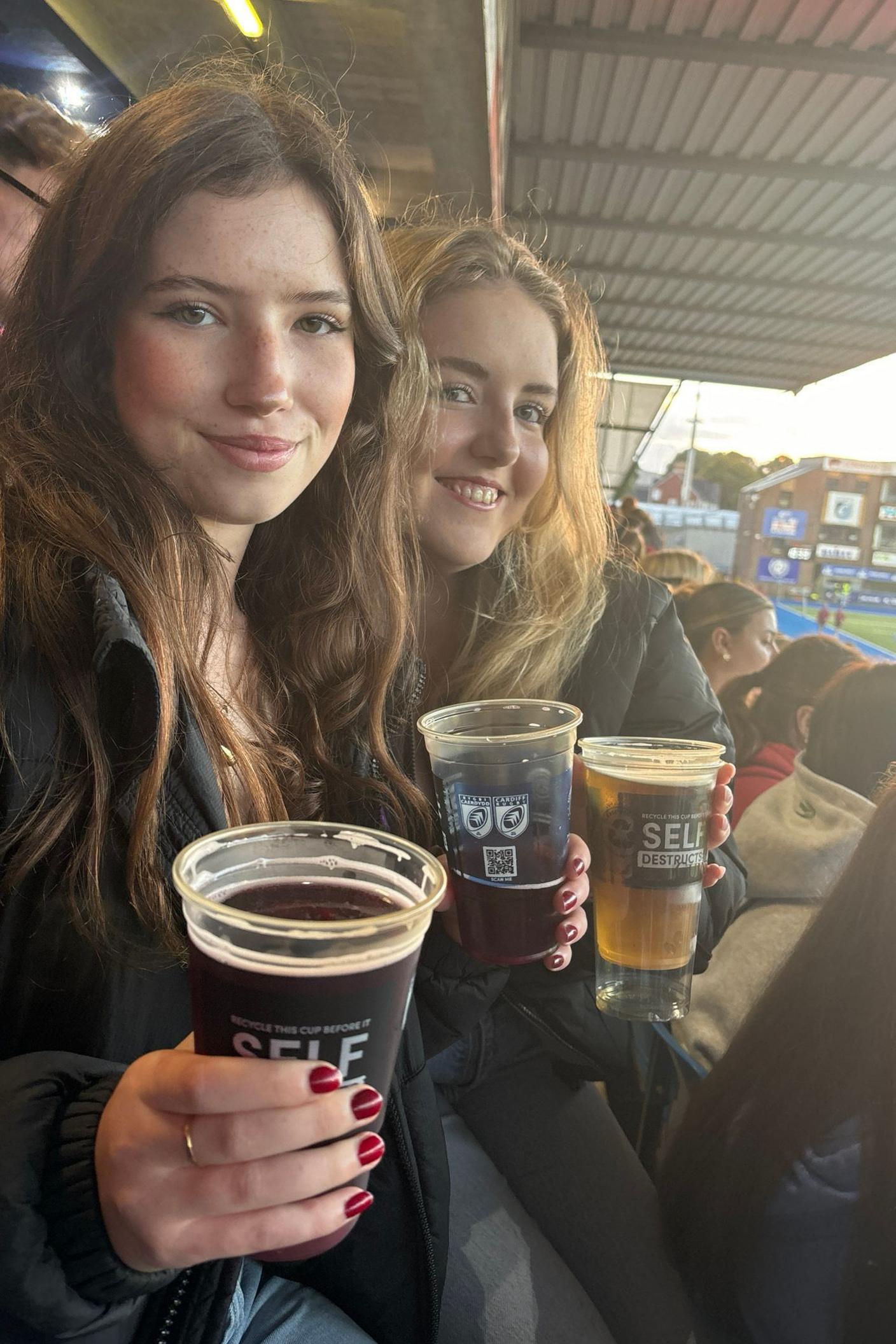 Two girls sat in the stands at a rugby stadium, smiling at the camera and holding pints. The one on the left is brunette, the other blonde, and they are both wearing black puffer jackets.