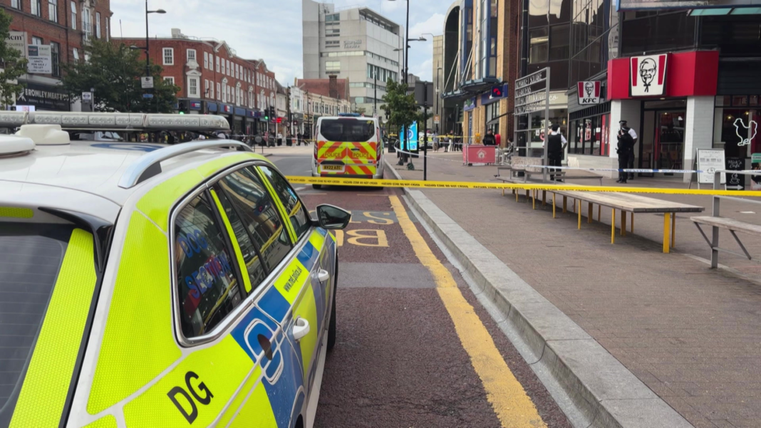 Police cordons block part of Bromley High Street as officers stand outside businesses near a KFC. A marked police car and police van are parked within the cordoned area, with the street largely empty behind the tape.