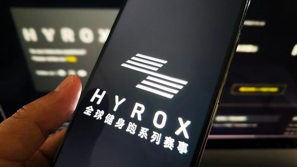 A woman holds a mobile phone with Hyrox branding on it.