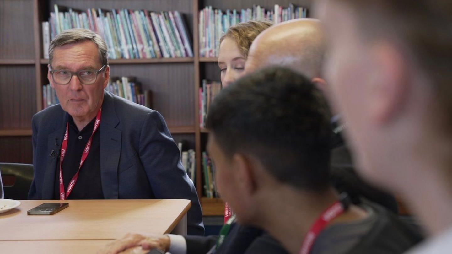 Alan Milburn author of the report sits on the left at the end of the table facing students in conversation, he wears a navy suit, red lanyard and black polo shirt. He looks serious in deep conversation.