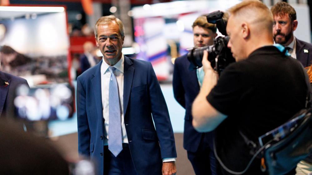Nigel Farage is wearing a blue suit, white shirt and blue patterned tie. A man with a camera is taking photos of him. Two other men can be seen in the background looking on.