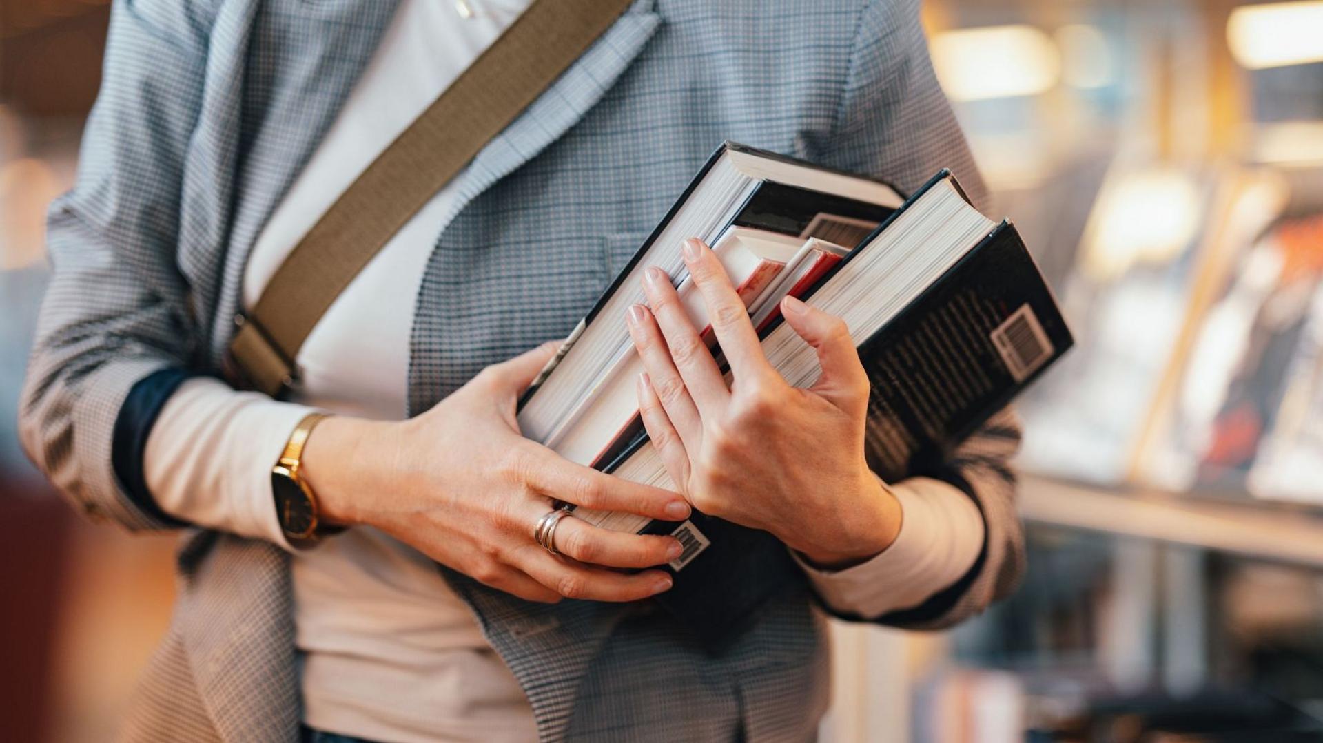 A person wearing silver rings and a gold watch carries an armful of books.
