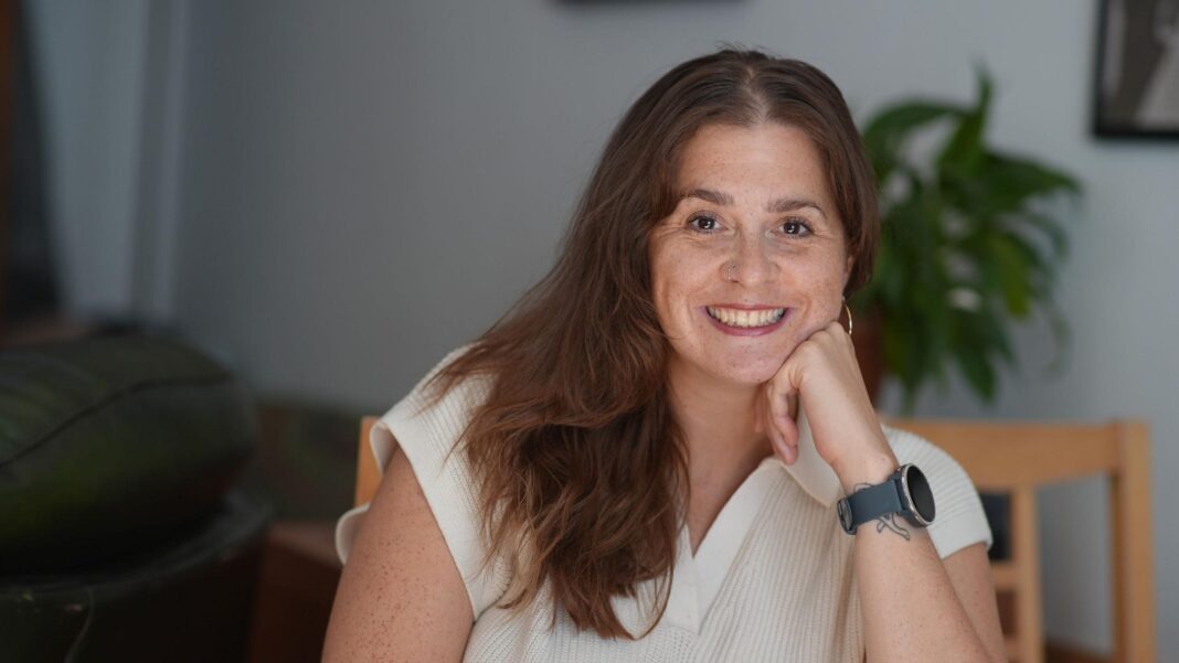 Jessica Hill, a woman in her 30s with long red-brown hair, smiles at the camera while resting her chin on her hand; she's wearing a white short-sleeved shirt with a collar, and a Garmin smartwatch on her wrist. She's sitting on a wooden chair, and a large house plant can be seen in the background, out of focus.