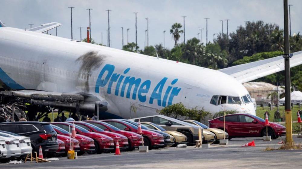 Tesla cars parked in a lot next to where a crashed Amazon Prime Air cargo plane came to rest