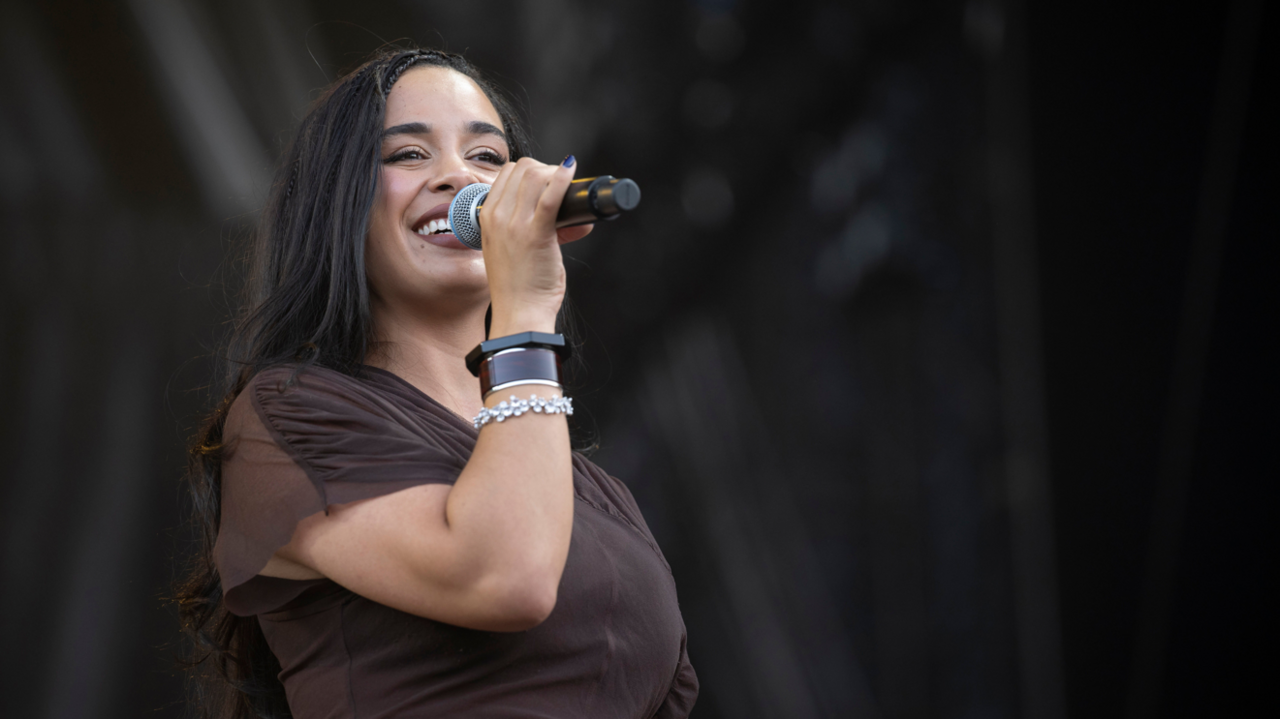 Person holding a microphone and performing on stage, wearing a dark brown outfit with long dark hair, photographed against a blurred black stage backdrop.