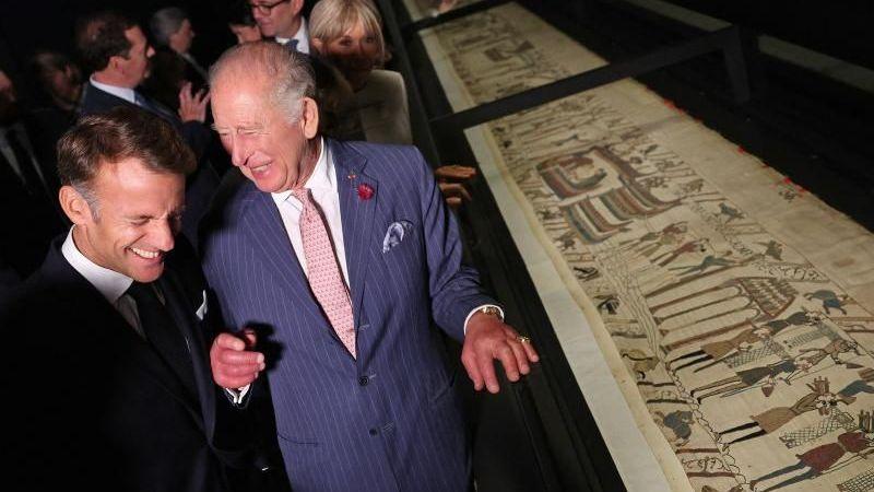 Emmanuel Macron and King Charles stand in front of the Bayeux Tapestry. They are both wearing blue suits with white shirts. Both are laughing.