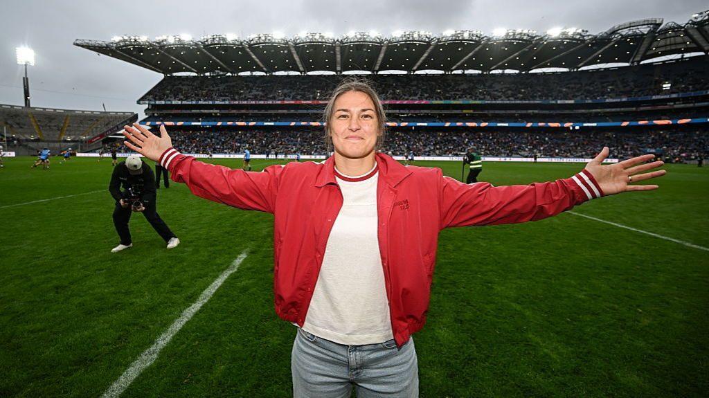 Katie Taylor is wearing a red jacket, white t-shirt and blue jeans and has her arm outstretched as she stands on the pitch at Croke Park