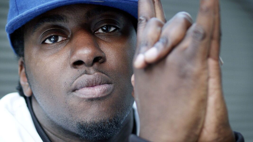 Sway wearing a blue cap and white shirt with his hands clasped in front of him