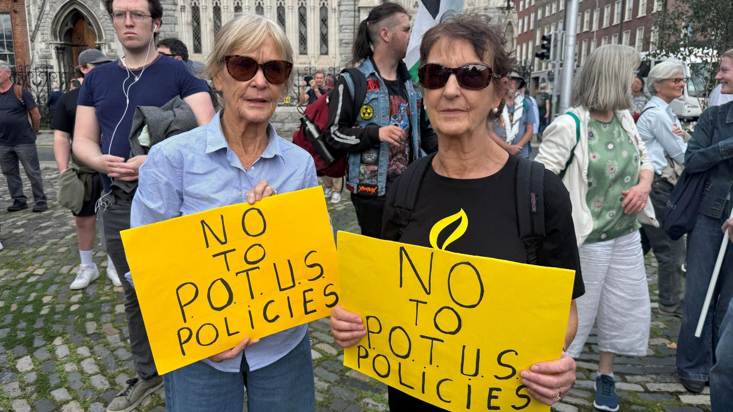 Woman with light hair, sunglasses, light blue shirt and jeans standing to the left with a yellow sign with black writing saying 'No to POTUS policies.' The woman on the right has short dark hair, sunglasses, black top and is holding the same sign.