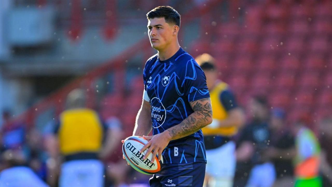 Louis Rees-Zammit stands on the pitch during a pre-game warm-up last season holding a rugby ball in two hands in front of him. He is wearing blue Bristol Bears kit