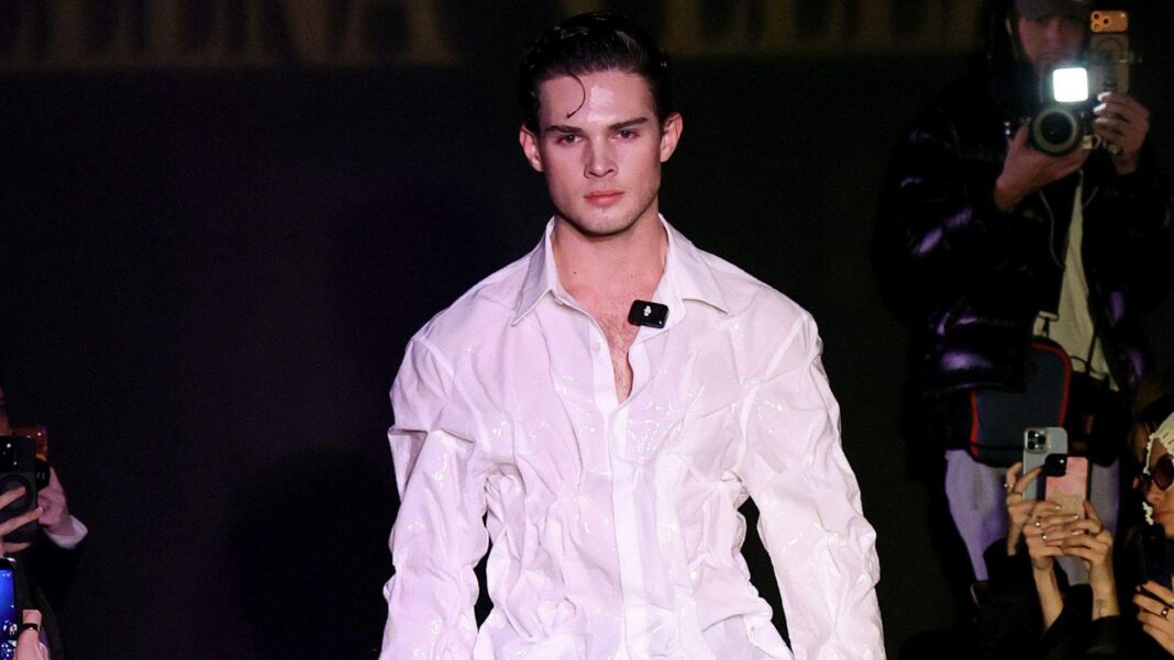 Clavicular walking down a fashion runway wearing a white shirt. He has dark hair.