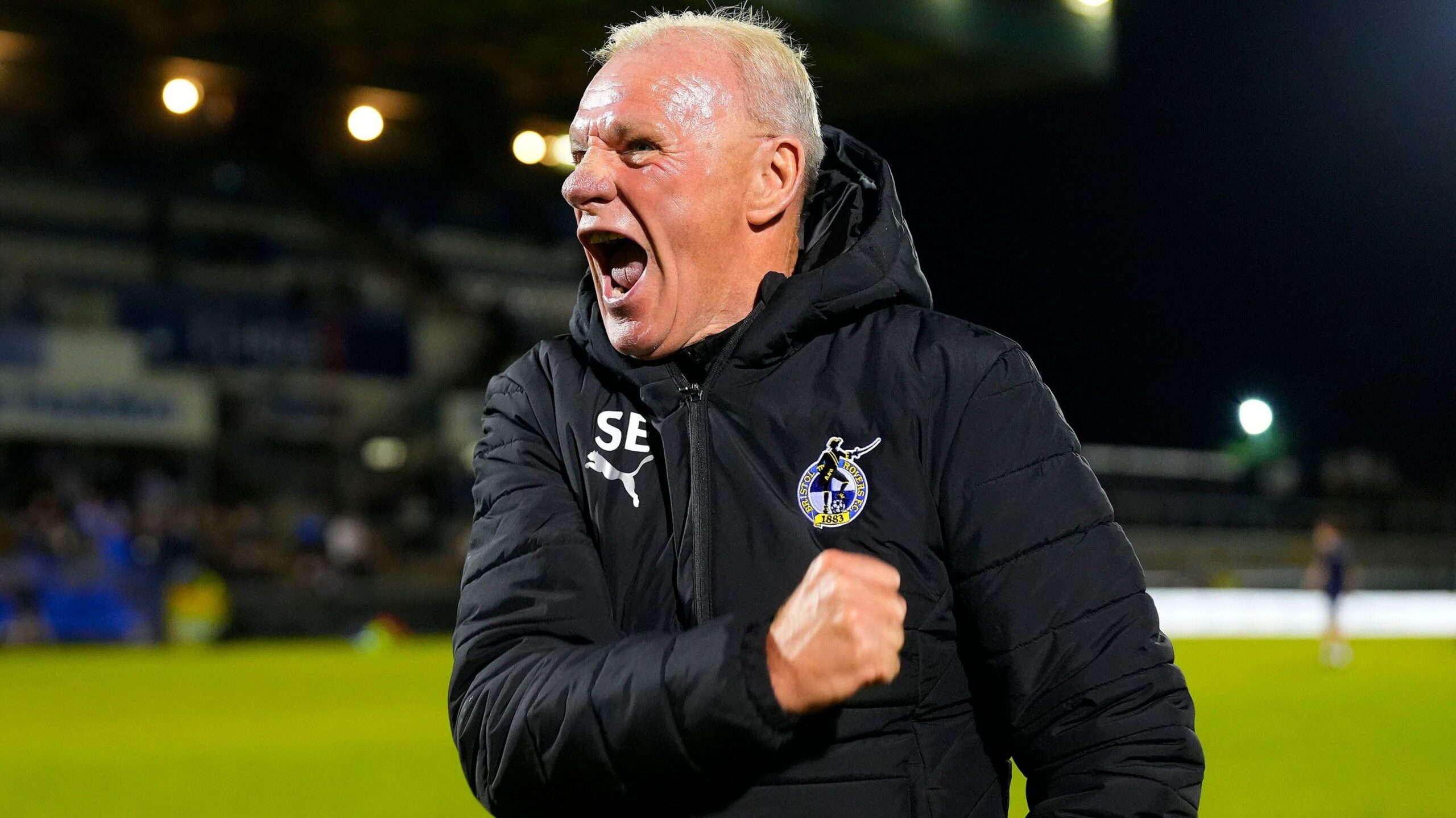 Steve Evans, wearing a black Bristol Rovers jacket, pumps his fists to celebrate the win against Colchester United