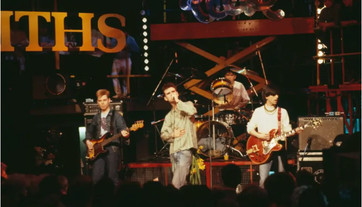 Four man stand on stage, one plays a red guitar, the other is singing into a microphone, the other is playing a bass guitar, and the other is playing drums. They are on a stage, with the front of a crowd staring back at them.