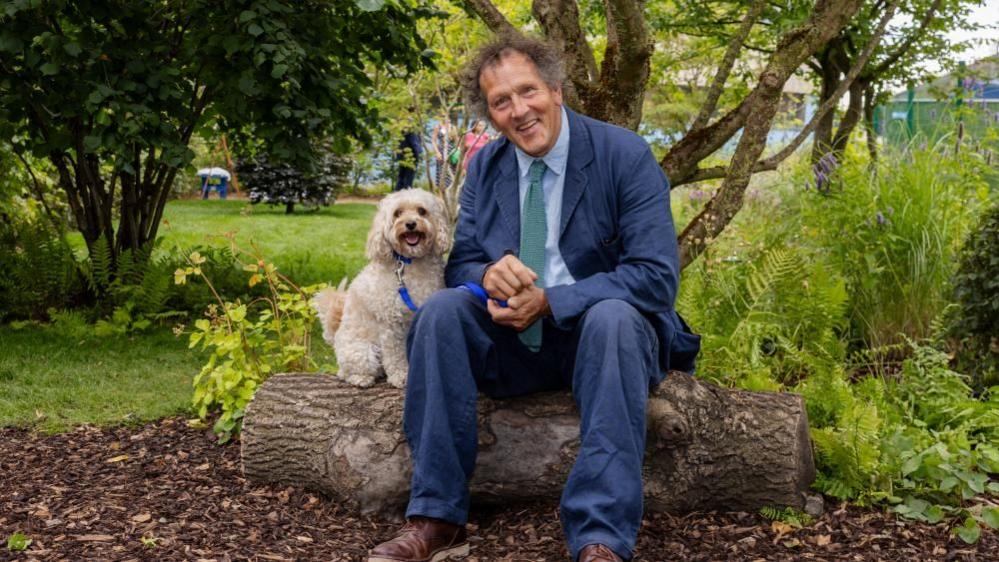 Don sits in a blue suit with a white curly haired medium sized dog. Both sit on a log and smile happily at the camera.