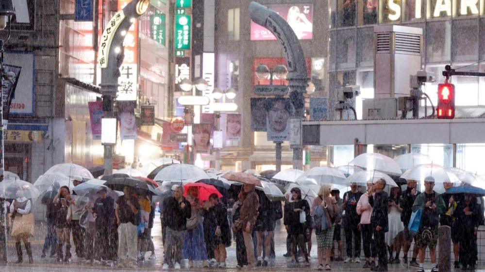 Several people in Japan wait to cross a road during a storm. They are all under umbrellas.
