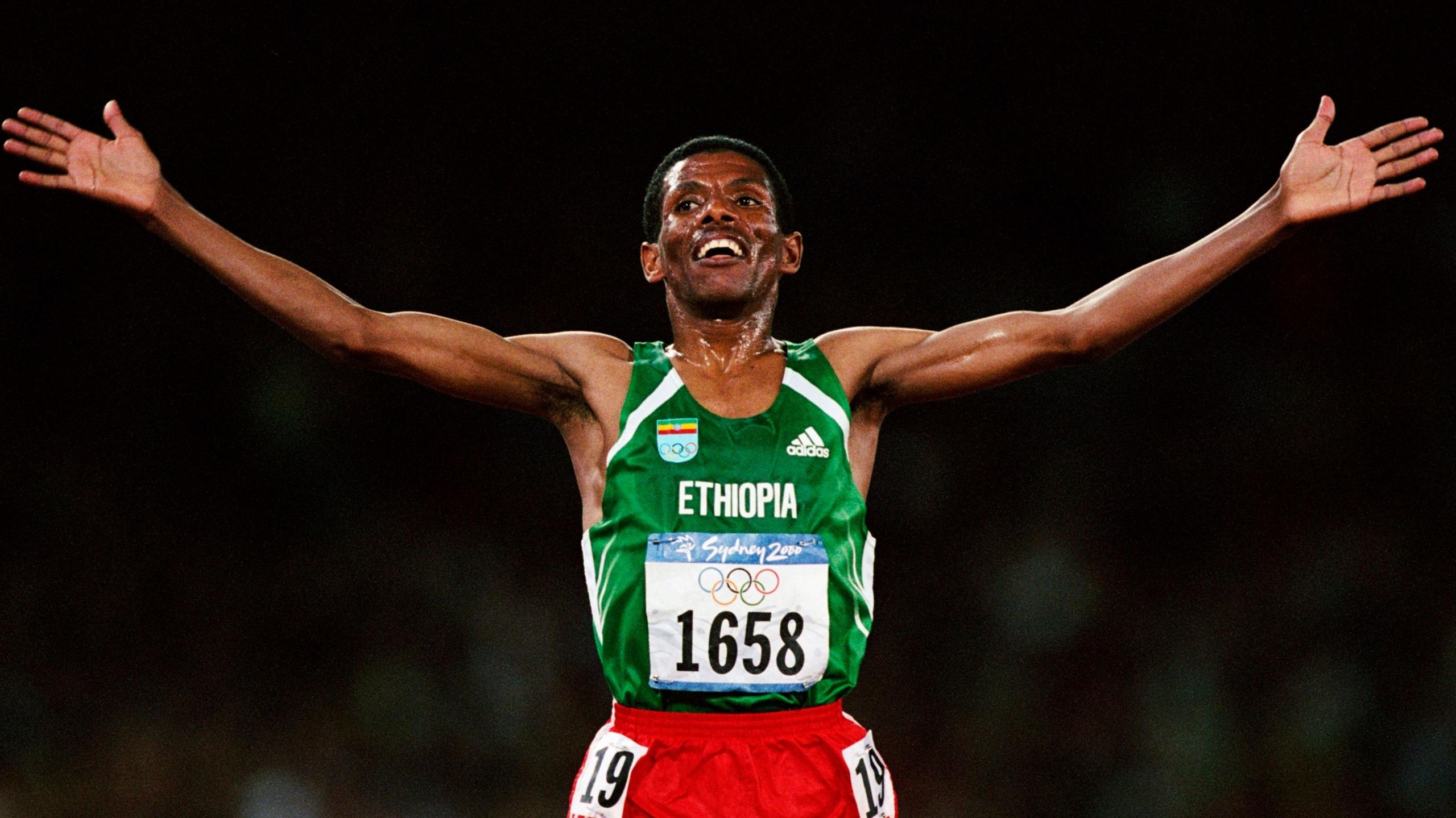 Haile Gebrselassie, wearing a green Ethiopia running vest and red shorts, holds out his arms in celebration as he smiles after winning the men's 10,000m race at the Sydney 2000 Olympic Games