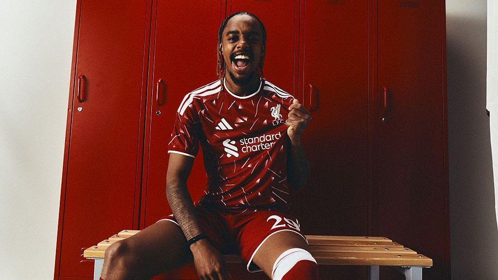 Bradley Barcola tugging on his Liverpool shirt badge with a smile on his face as he celebrates joining Liverpool