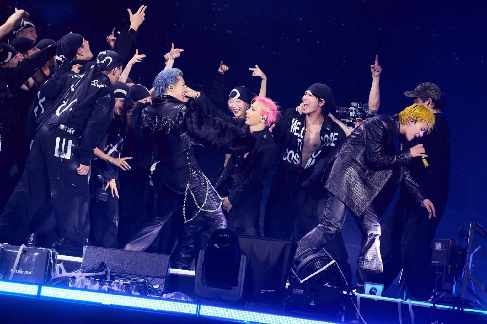 A performer with bright yellow hair leans forward at the front of a stage, holding a handheld microphone. Around the performer, a group of performers and dancers in black outfits gather in a tight formation, with several raising their arms. Another performer with blue hair and a black outfit stands near the centre, while a performer with pink hair is visible beside them. They are illuminated by blue lighting against a dark background.