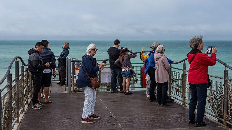 Group of people standing on a pier looking out to sea. Many are wearing coats, and it also looks quite breezy