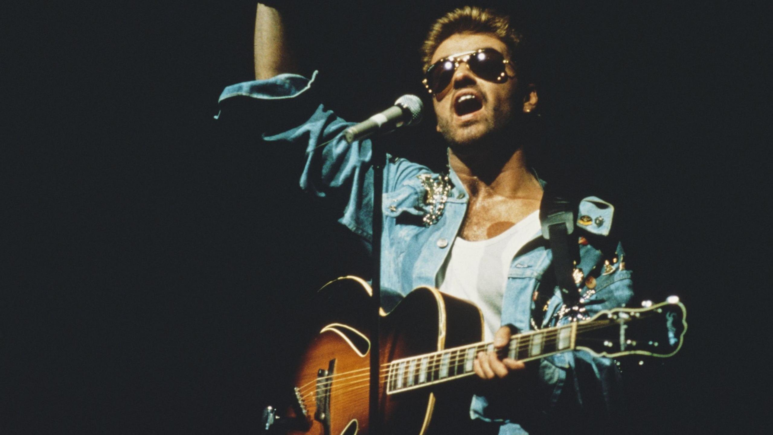 George Michael performing on stage with an acoustic sunburst guitar, and wearing aviator-style Rayban sunglasses.