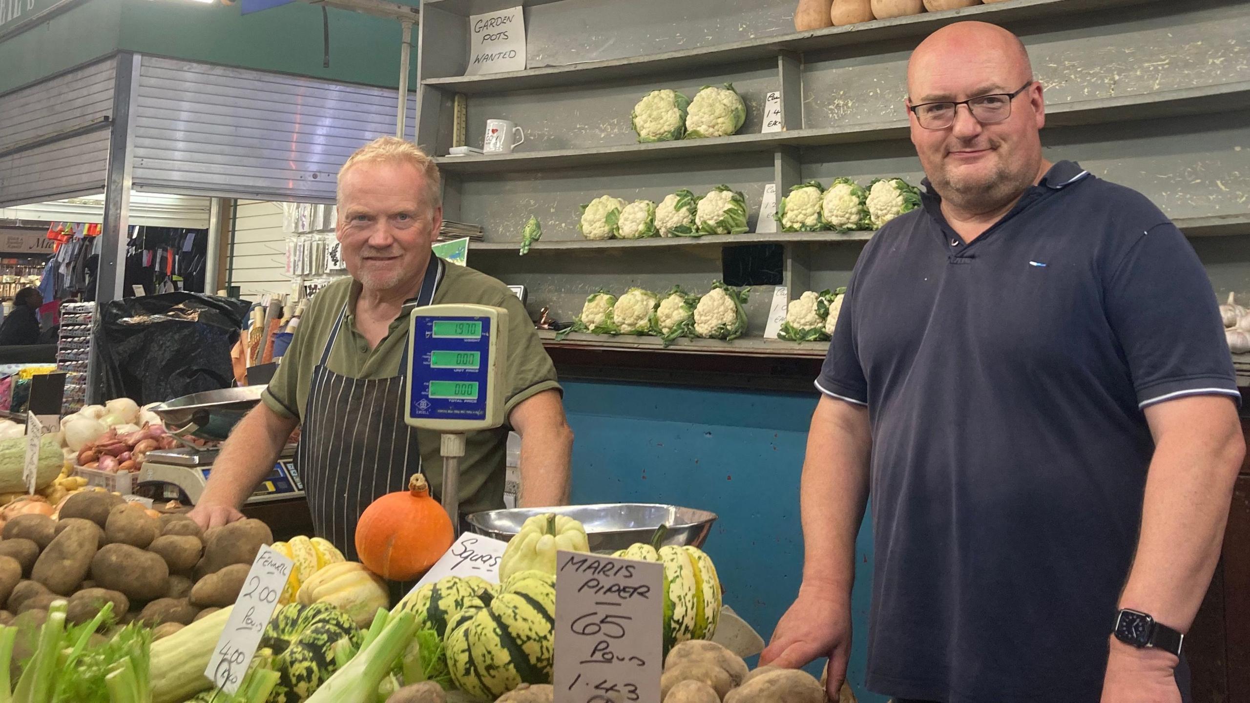 Two people stand behind a traditional market produce stall surrounded by fresh vegetables. In the foreground are potatoes, striped squashes, pumpkins, celery and onions, while rows of cauliflowers are displayed on shelves behind the counter. Handwritten price signs are attached to some of the produce, and parts of the indoor market can be seen in the background.