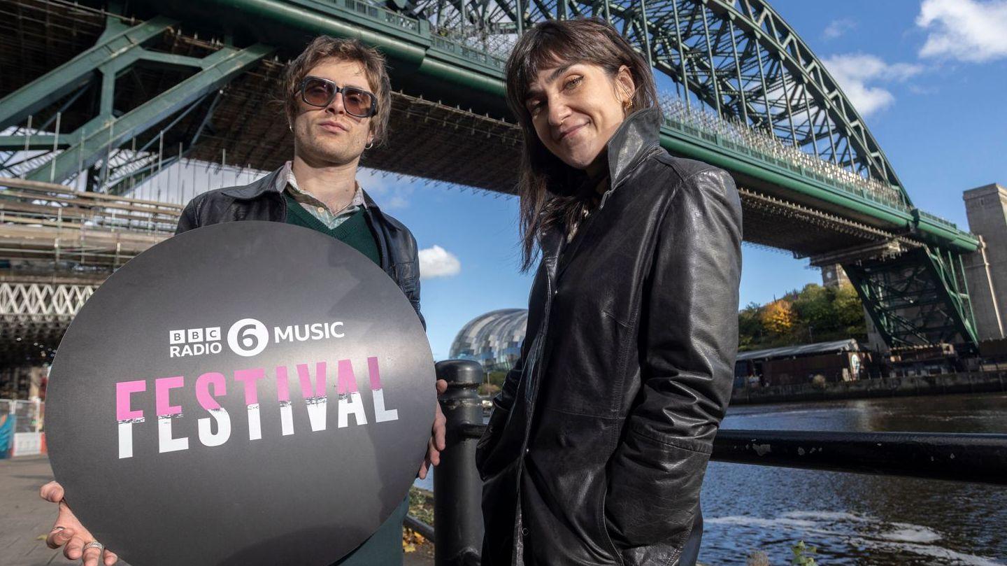 6 Music presenters Nathan Shepherd and Deb Grant standing below the Tyne Bridge which is covered by some scaffolding. The camera is slightly below them and they are looking down. Shepherd is holding a black circular sign which reads BBC Radio 6 Music Festival in pink and white lettering. He has brown, short hair and is wearing large glasses, a black leather jacket on top of a green jumper and shirt. Grant is on the right and has long, dark hair with a full fringe. She is smiling and standing slightly on an angle and wearing a black leather jacket with her hands in the pockets. The River Tyne is on the right and look calm. The Glasshouse can just be seen in between the presenters.