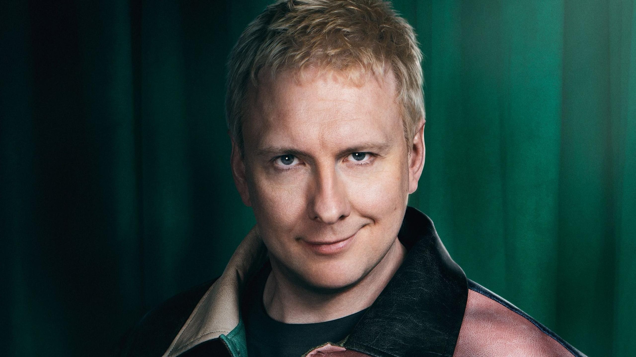 Joe Lycett has short blonde hair and wears a mulit coloured jacket and smiles mischievously at camera