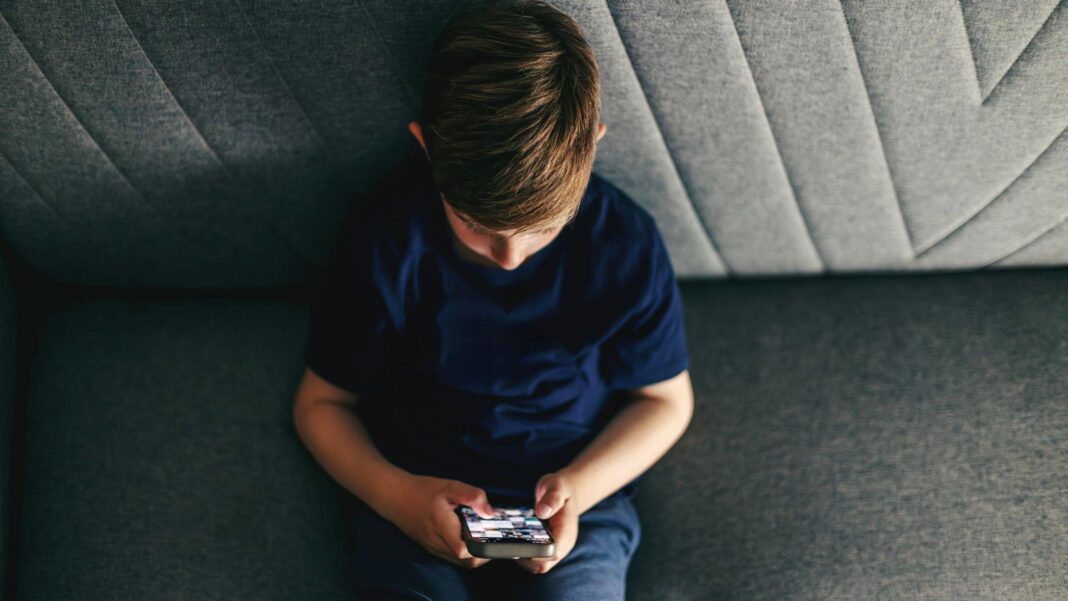 Young boy sat on grey couch holding smartphone.