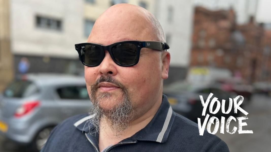 ‘People call them pervert glasses’: Blind users fear backlash over smart tech Portrait of Fraser Fleming wearing dark sunglasses and a navy zip-neck polo shirt, standing on a wet city street with parked cars and buildings blurred in the background.