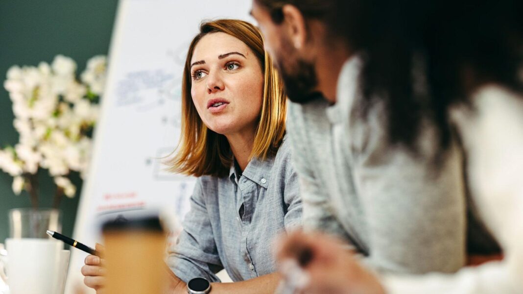 A woman speaking is in a meeting with a whiteboard in the background. Some other people including men are blurred n the foreground