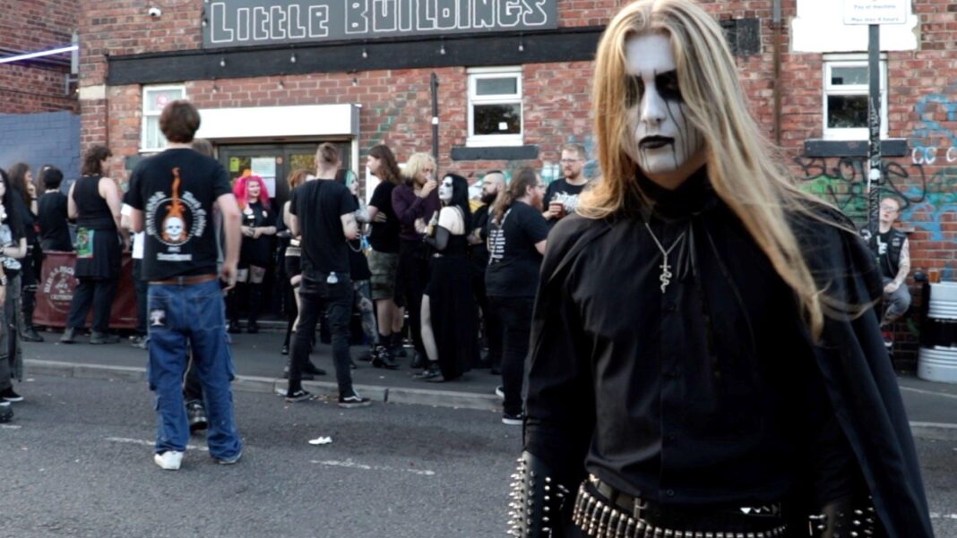 Black metal mourns pioneering Venom frontman Envus is stood in front of a crowd outdoors. He is wearing all black, has long brown hair and his face painted white with black accents.