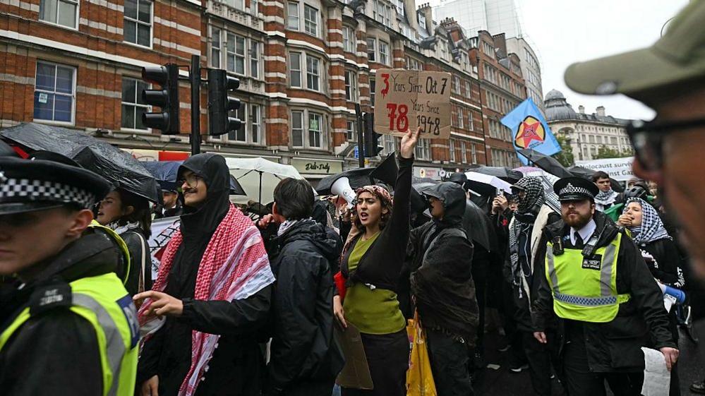 A group of people, some waving flags or holding placards, take part in a pro-Palestinian march in London. One protester can be seen holding a sign that reads "3 years since October 7, 78 years since 1948". Police can be seen escorting the march.