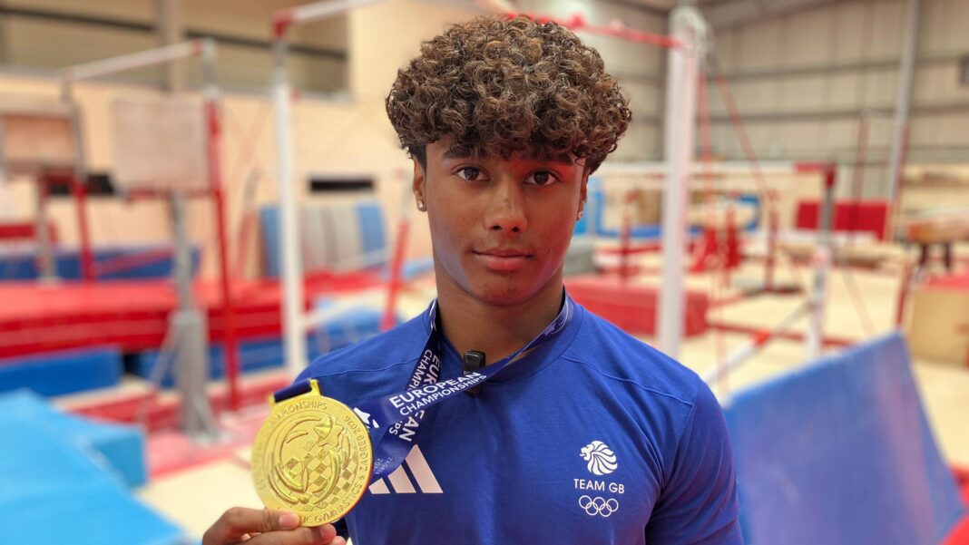 GB stars aim for Olympic spots at World Championships – live on BBC Uzair Chowdhury looking at the camera while clutching his gold medal. He is wearing his blue Team GB uniform and is stood at his gym, with crash mats and equipment blurred out behind. Chowdhury has curly brown hair and is wearing stud earrings.