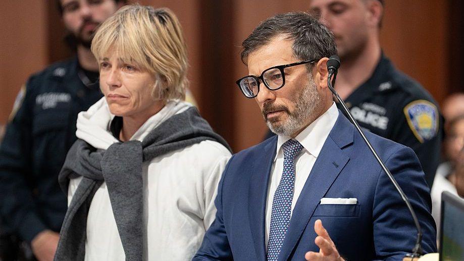 Two people in a courtroom, the woman to the left has short cropped blonde hair, a sad expression and is wearing a white hoodie. The man on the right has dark hair, a scruffy beard, and is wearing a blue blazer.