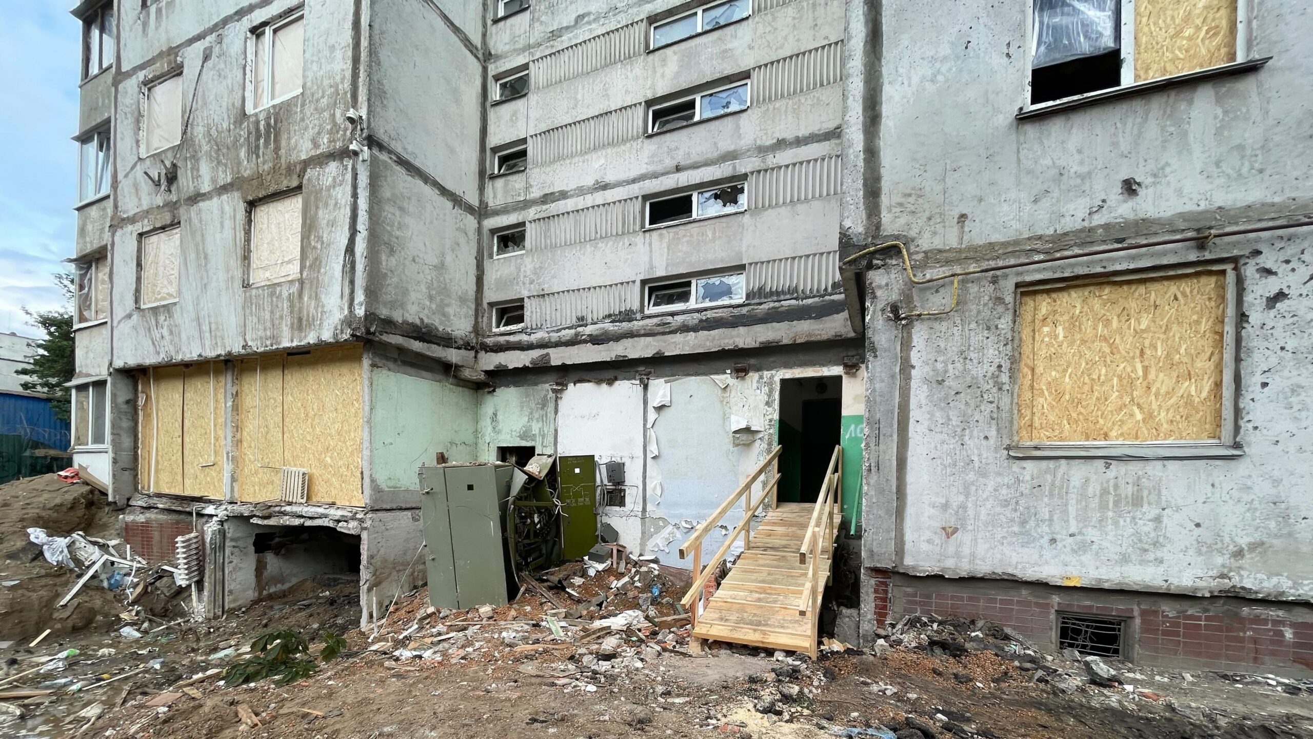 Photo shows the lower floors of an apartment block with multiple windows blown out and boarded up. Ground around the block is muddy and damaged.
