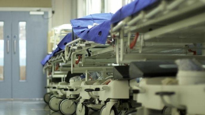 A row of empty hospital trolleys line a brightly lit corridor, their wheels reflecting on the polished floor. Blue protective sheets cover the beds, while there are closed double doors at the far end