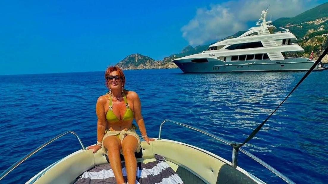 Kershaw is sat down on the front of a boat facing away from the sea. Behind her is a large yacht and a coastline.