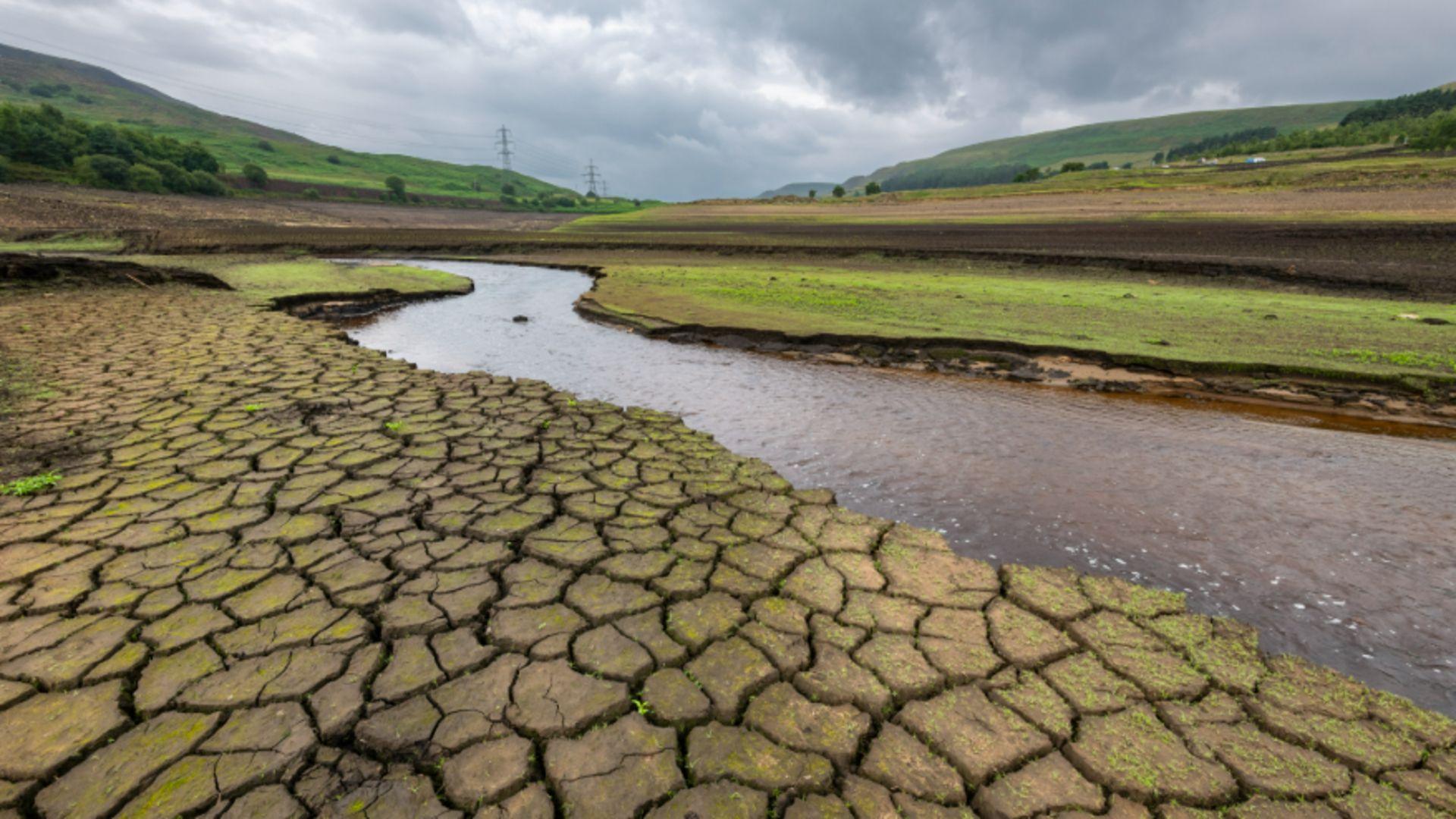 An image of drought affected ground alongside a water stream, with green hills in the background.