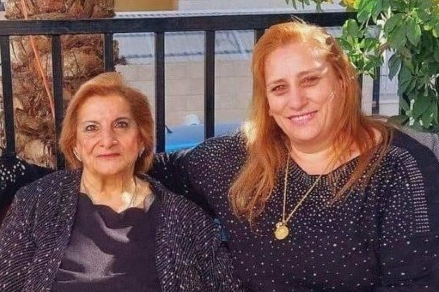 Nahida and Samar Anton sit smiling next to each other in front of a railing. They are both wearing black blouses with small white spots while Samar wears a gold chain with a gold pendant.