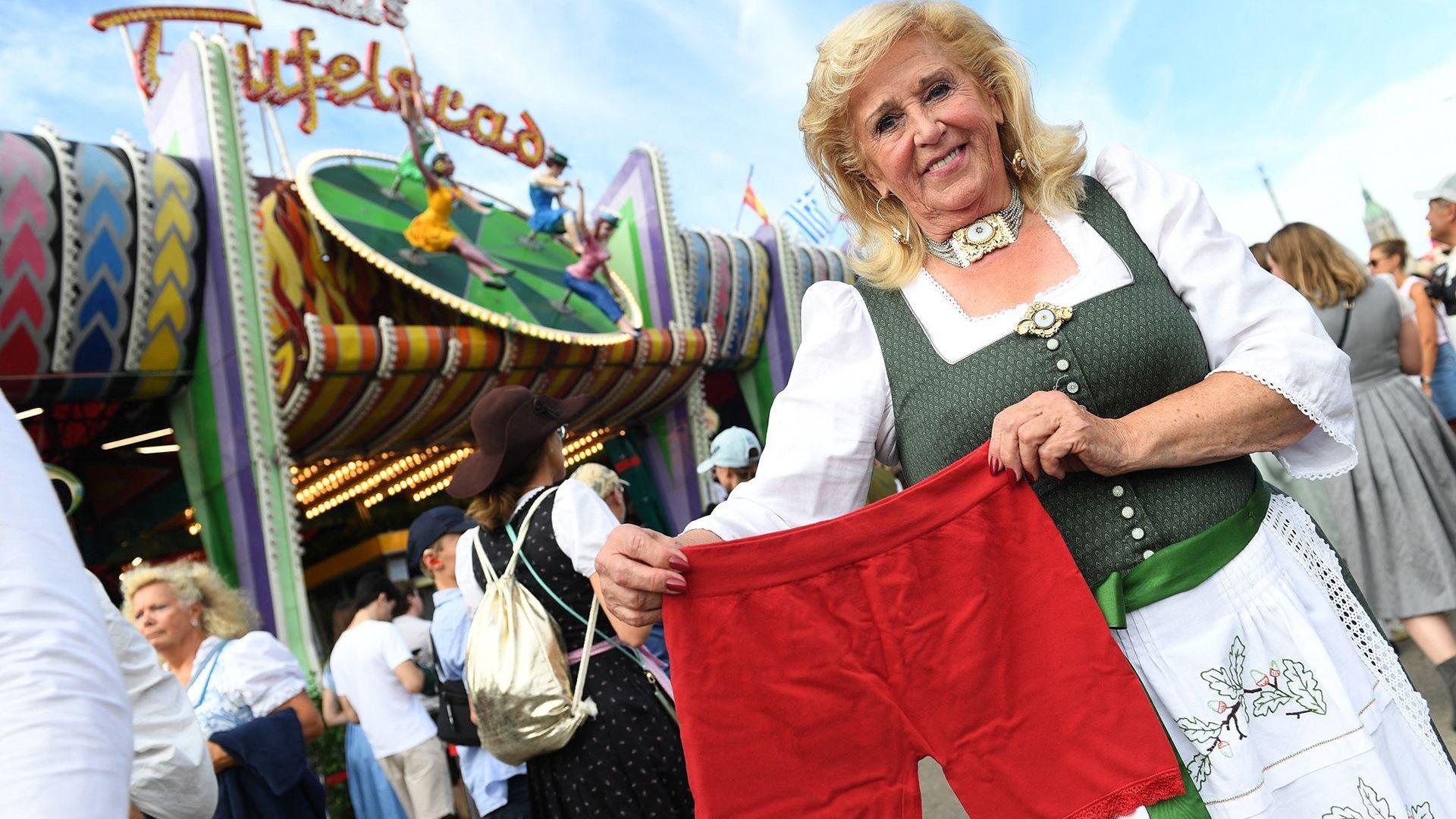 Elisabeth "Lisi" Polaczy, the operator of the Teufelsrad, stands in front of the traditional "Teufelsrad" ride. She is holding a red pair of cycling shorts while wearing a green and white traditional German dress.