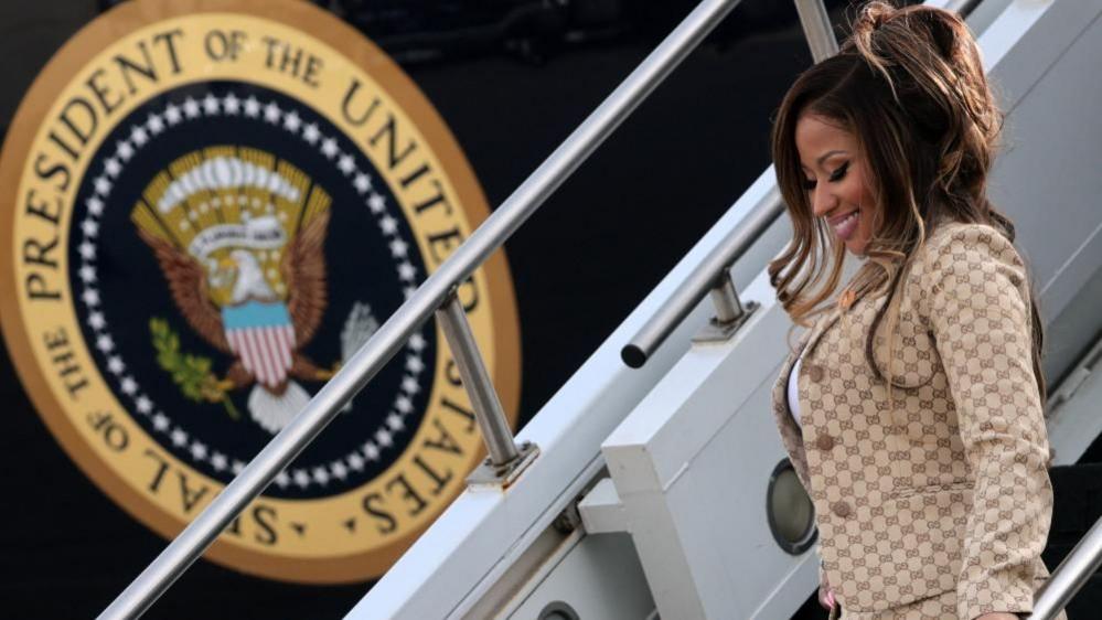 Minaj, smiling in her gold suit, walks down the steps of Air Force one, with the plane's livery clear in the background.