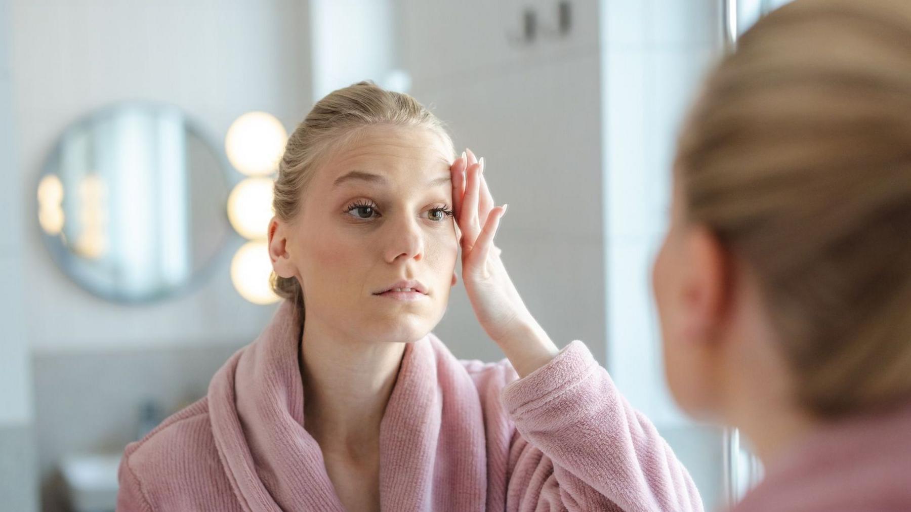 A tired woman wearing a pink dressing gown looks at herself in the mirror, with her hand touching the side of her forehead