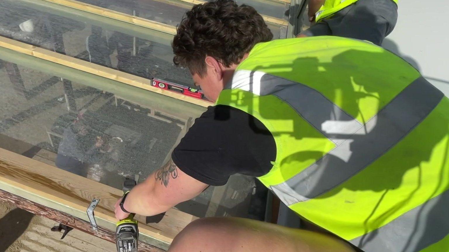 A man with black curly hair uses a drill on a piece of wood on a glass panel. He is wearing shorts, a black T-shirt and a yellow high-vis jacket. He has a tattoo on his left arm and has his back to the camera.