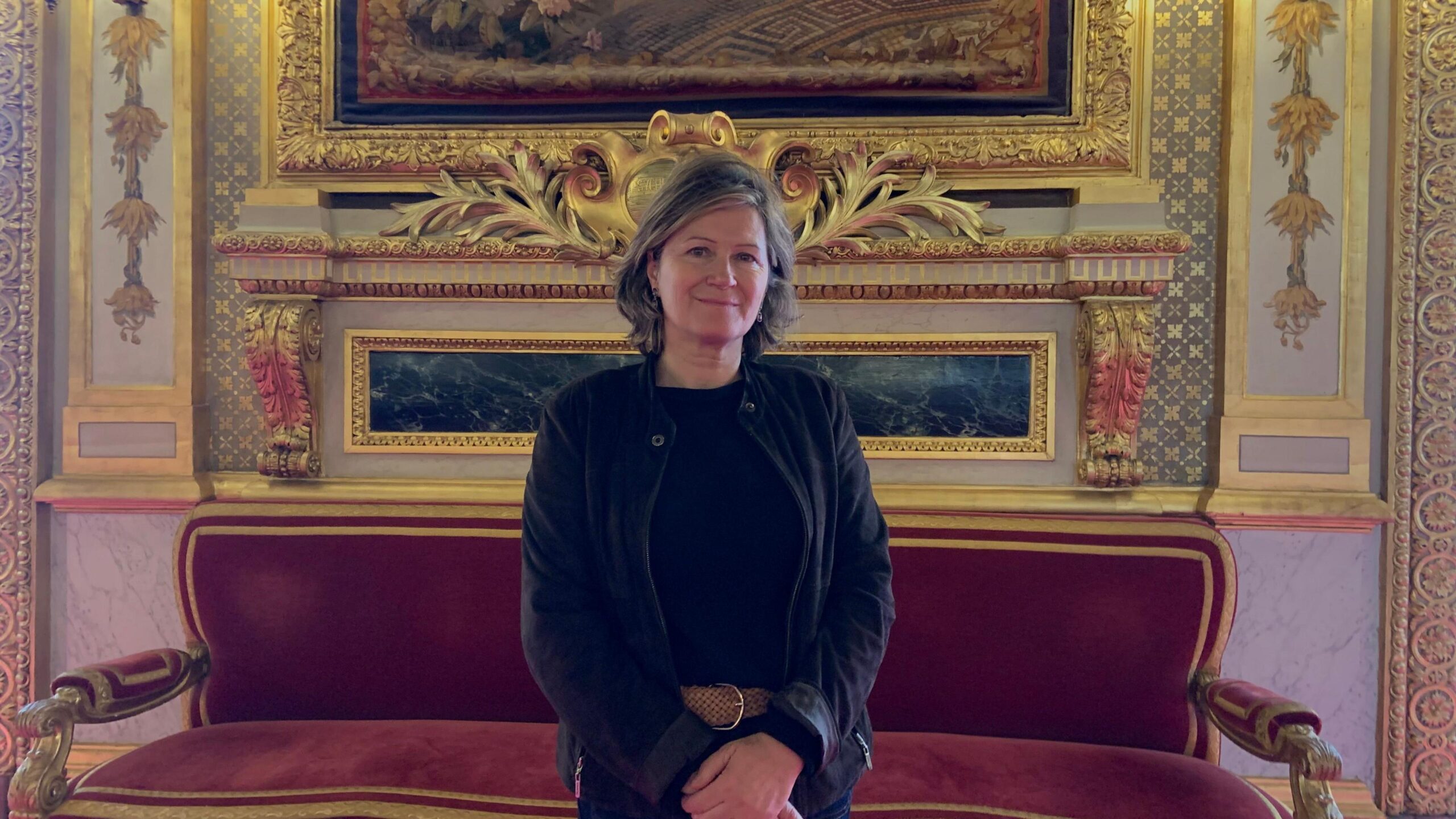 A woman with a dark top and cardigan and greying hair stands in front of an ornate setting with gilded decorations on the wall