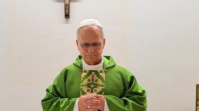 Pope Leo celebrates mass from hospital, wearing a green robe and standing before a white wall adorned with a wooden crucifix.