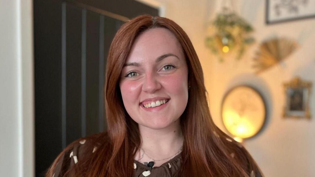 ‘Ominous signs’ of winter pressures as NHS waiting list grows Amy-Jane is stood in her living room and is smiling at the camera. She has long reddish-brown, straight hair and is wearing a brown and white spotty blouse.