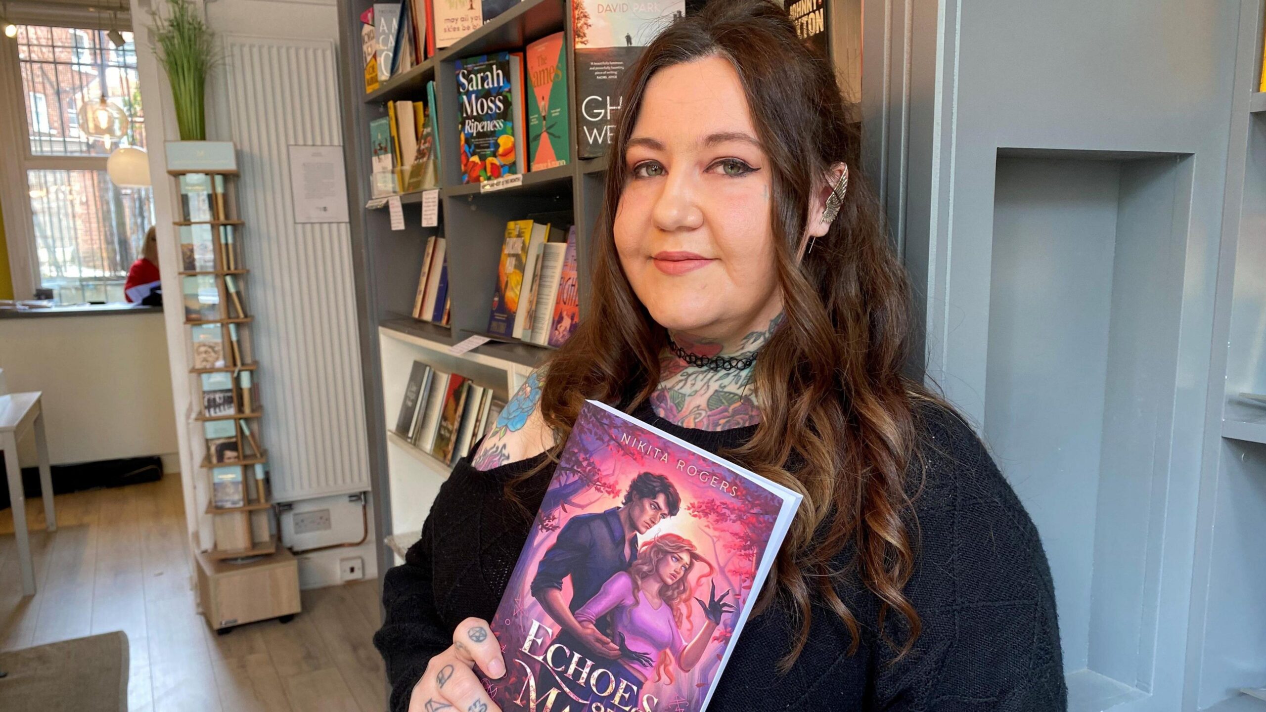 Nikita has long curled brown hair, she is wearing a black shoulder and holding a book which depicts a a brown haired man behind a blonde haired lady. Nikita has tattoos on her hand, shoulder and neck. in the background grey shelves with books can be seen.