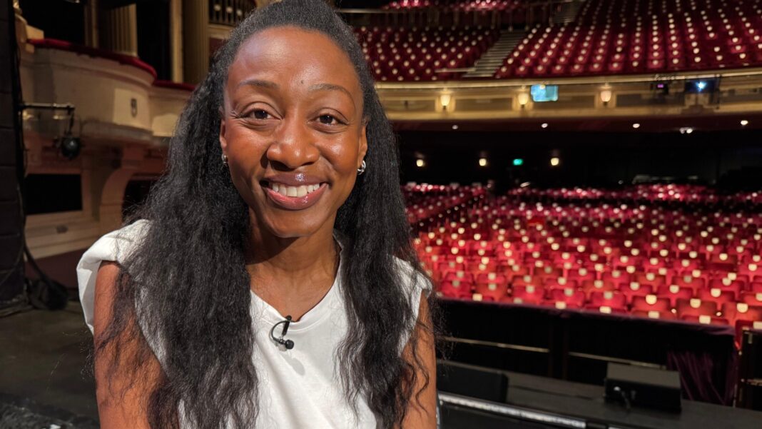 Suffragette role ‘a gift’ says Beverley Knight A woman wearing a white top and a clip-on microphone sits on the stage at a theatre, with rows of red auditorium seats visible in the background.